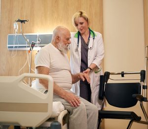 Am Foto ist ein männlicher Patient, sitzend am Krankenhausbett, mit seiner Ärztin zu sehen.