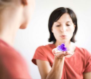 Am Bild ist eine Frau im roten T-Shirt zu sehen. Sie hält eine violette Blüte in ihrer rechen Hand und versucht diese zu verblasen.